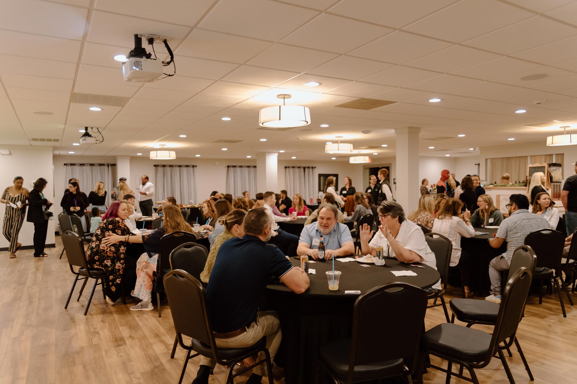 People sitting at round tables in a well-lit room, talking and socializing at an indoor event.