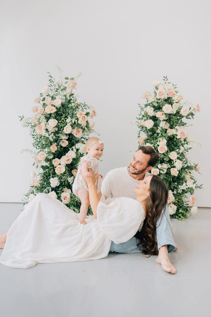A family with a baby sits and smiles together in front of two tall floral arrangements with pale roses.
