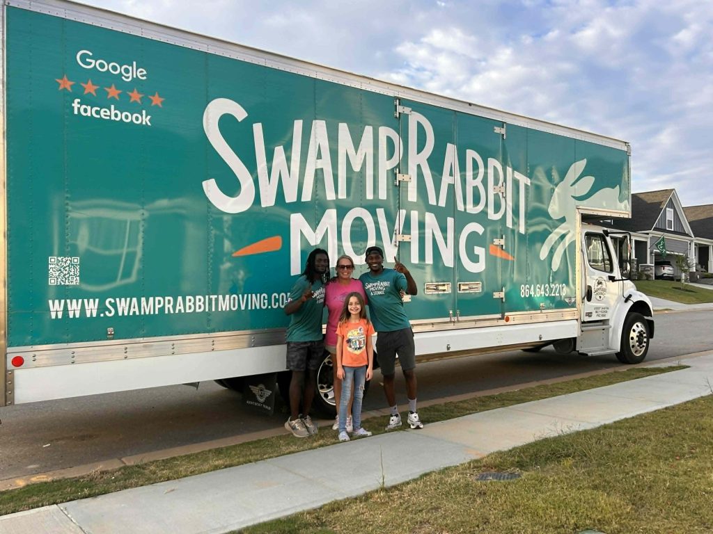 Four people stand smiling in front of a large Swamp Rabbit Moving truck parked on a suburban street.