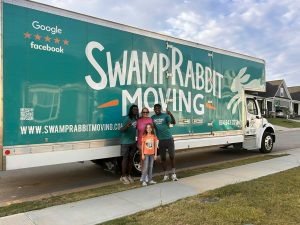 Four people stand smiling in front of a large Swamp Rabbit Moving truck parked on a suburban street.