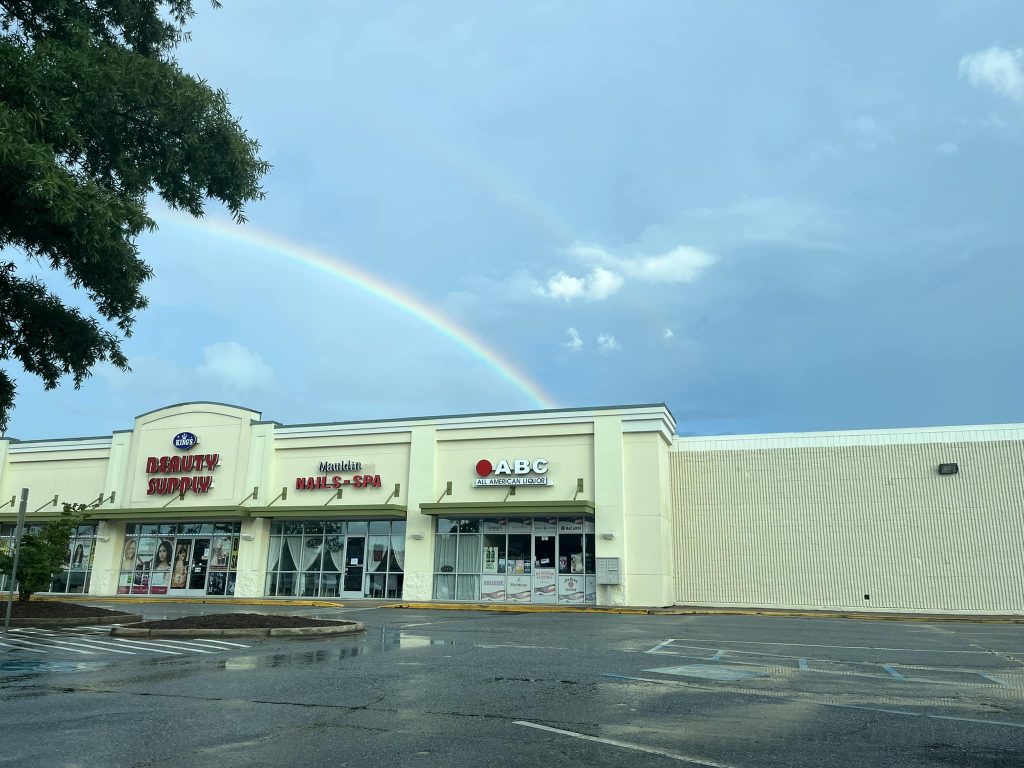 A faint double rainbow appears in the sky above a strip mall with empty parking spaces.