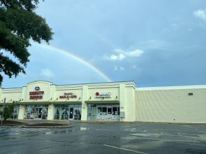 A faint double rainbow appears in the sky above a strip mall with empty parking spaces.