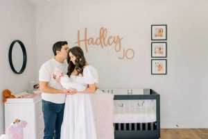 A couple lovingly holds their baby in a nursery with "Hadley Jo" on the wall above a crib.