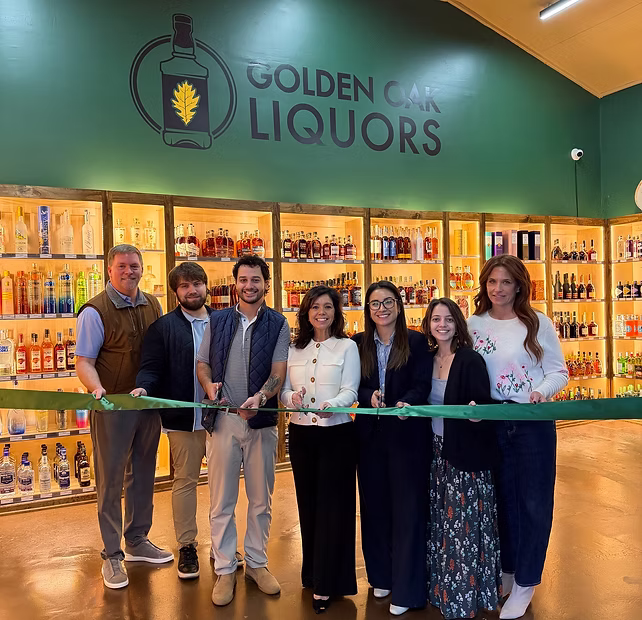 Seven people stand in front of liquor shelves, cutting a green ribbon at Golden Oak Liquors.