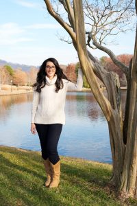 Woman in a white sweater and boots stands by a tree near a lake on a sunny day.