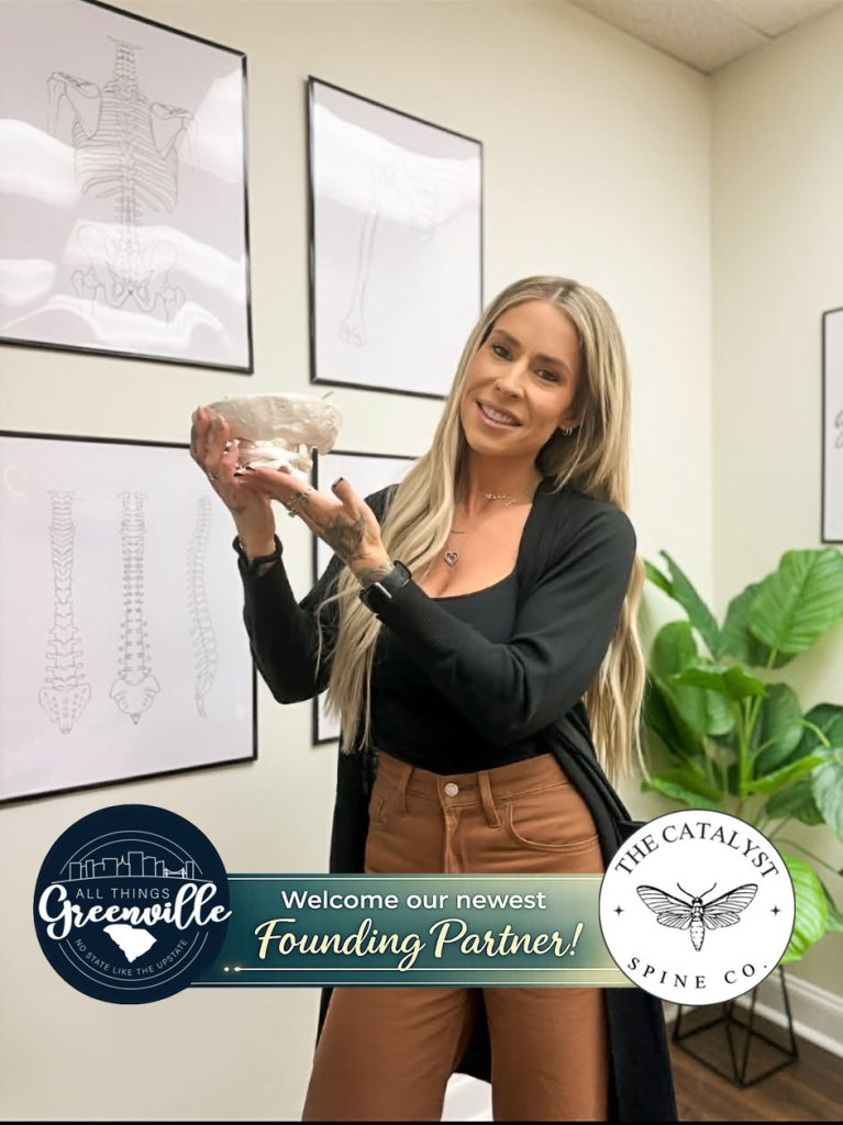 A woman smiles holding a spine model, standing in an office with spine diagrams on the wall.