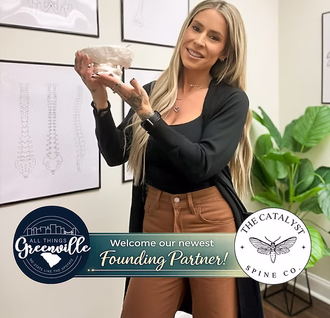 A woman smiling and holding a spine model in an office with spinal diagrams on the wall.