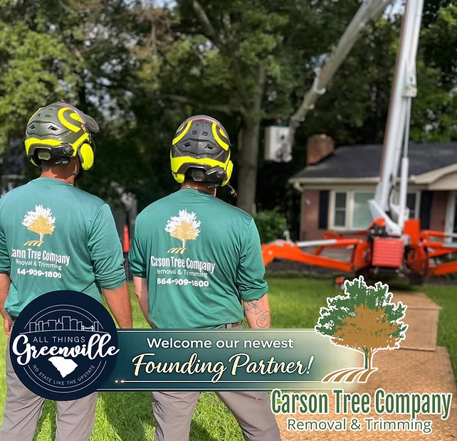 Two workers in green shirts and helmets stand by tree trimming equipment in a residential yard.
