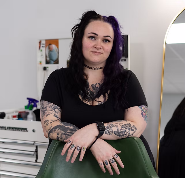 Woman with dark hair and tattoos leans on a green chair in a modern, bright room with salon equipment.