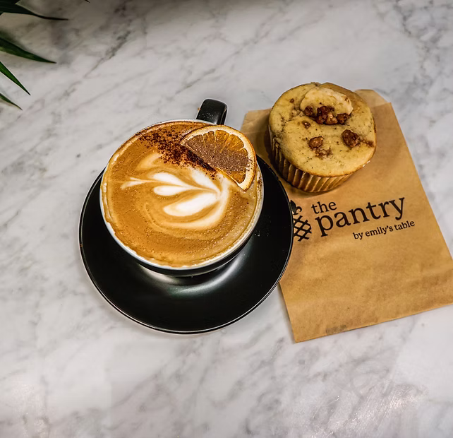 A latte with latte art and an orange slice, next to a muffin on a "the pantry by emily’s table" paper bag.