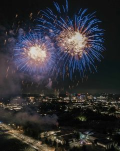 Colorful fireworks burst in the night sky above a brightly lit cityscape, with smoke drifting below.