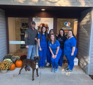 Six people in blue and black scrubs and a dog pose outside a veterinary clinic decorated with pumpkins and flowers.