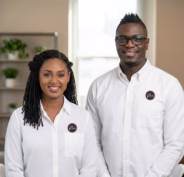 Two people in white shirts with matching round logos, smiling and standing indoors in a bright room.