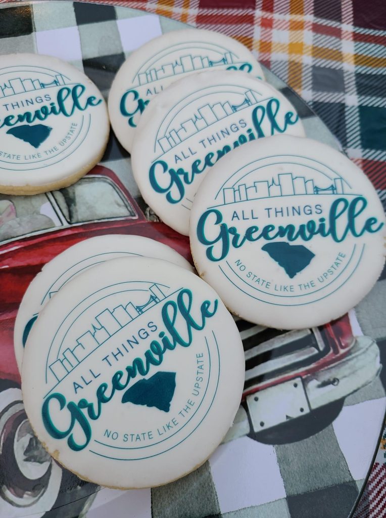 Round cookies with "All Things Greenville" and a city skyline printed on them, placed on a plaid cloth.
