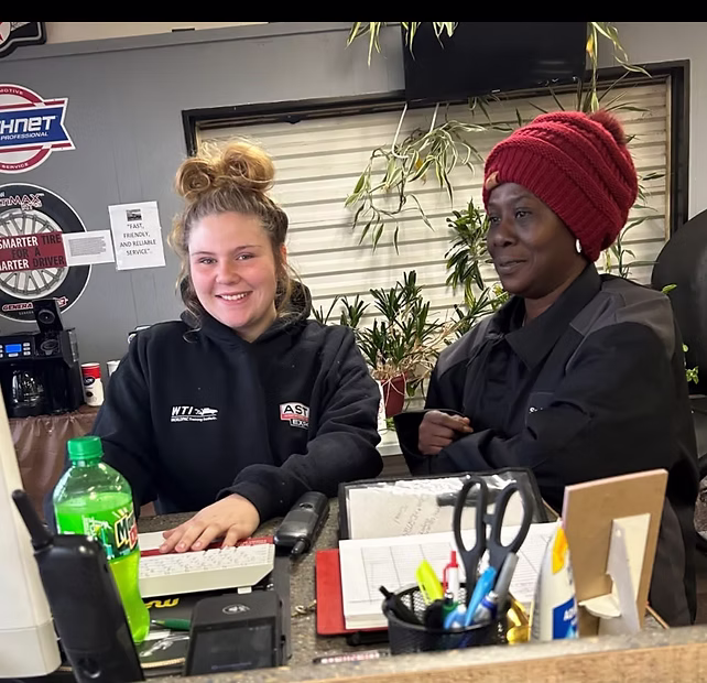 Two women smile behind a counter with a keyboard, papers, and a bottle of Sprite visible in front of them.