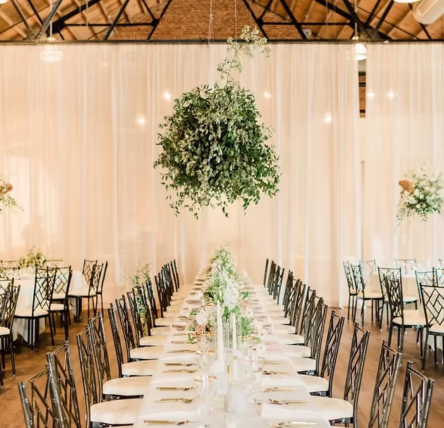 Elegant banquet hall with long dining tables, black chairs, and a hanging green floral arrangement centerpiece.