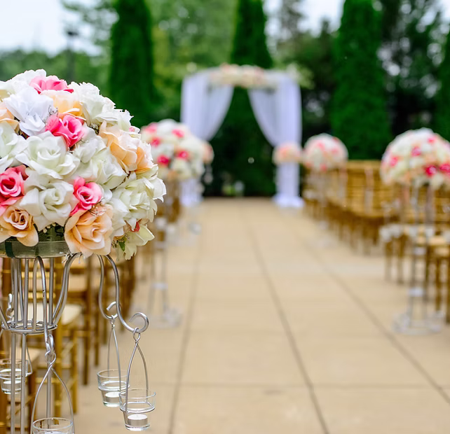Outdoor wedding aisle lined with flower arrangements and chairs, leading to a decorated archway.