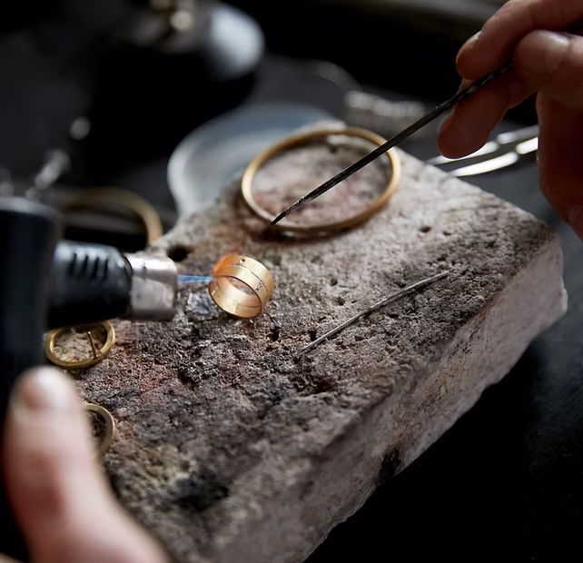 Close-up of hands soldering a gold ring with a torch on a workbench with jewelry tools and a stone block.