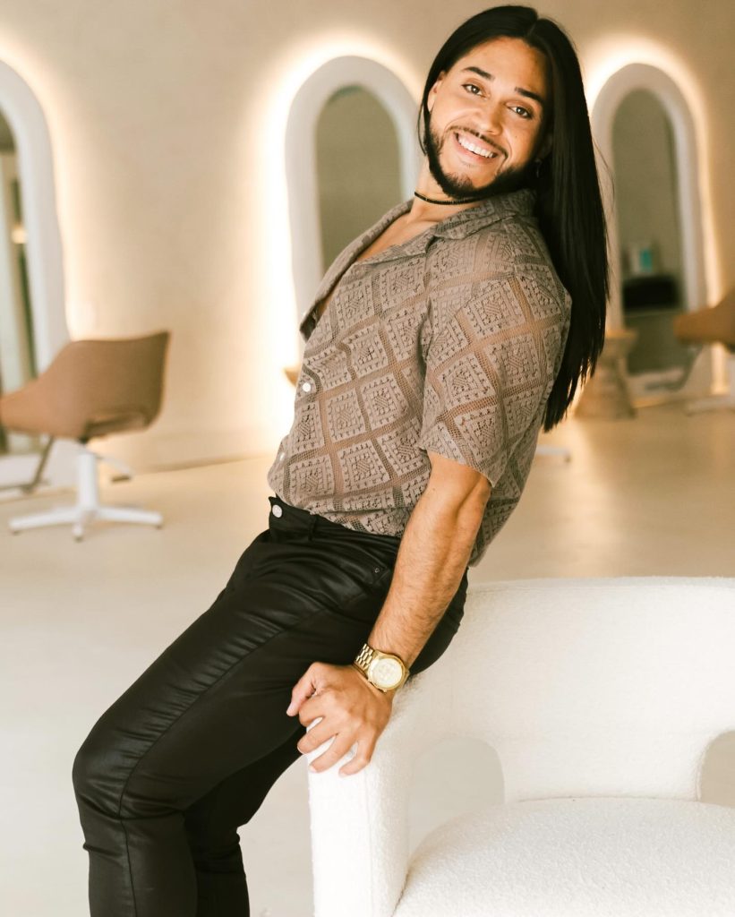 Smiling person with long hair poses on a white chair in a modern, bright salon with arched mirrors.