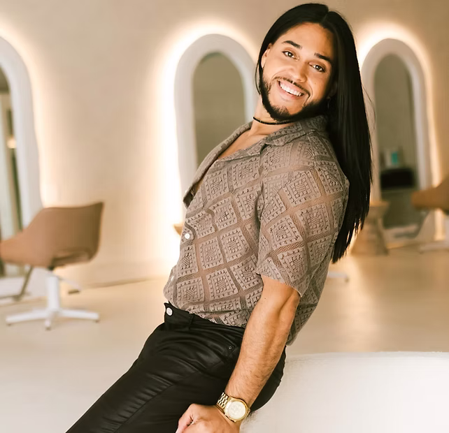Smiling person with long dark hair poses indoors, wearing a patterned shirt and black pants.