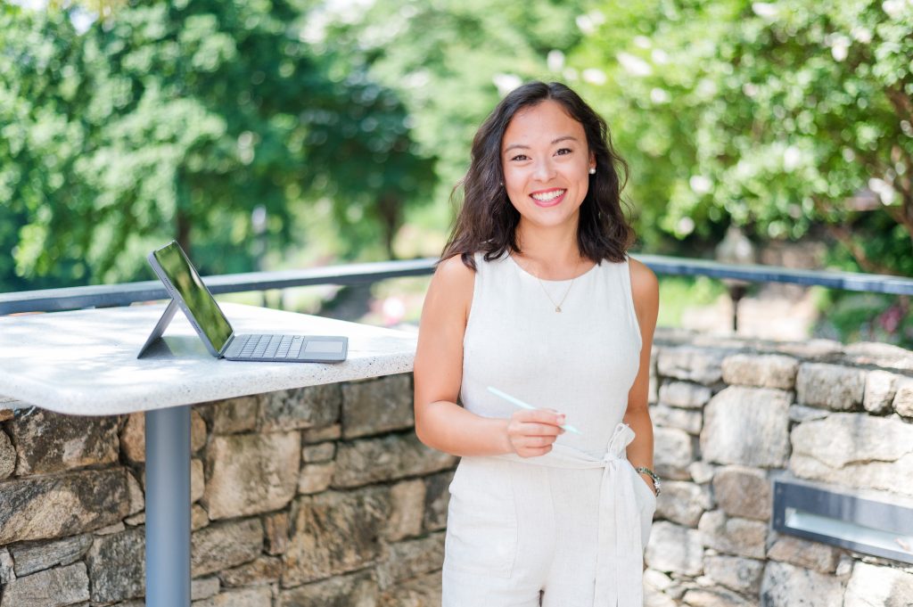Woman in a light outfit smiles outdoors by a stone wall and a table with a tablet and keyboard on it.