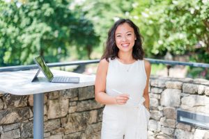 Woman in a light outfit smiles outdoors by a stone wall and a table with a tablet and keyboard on it.