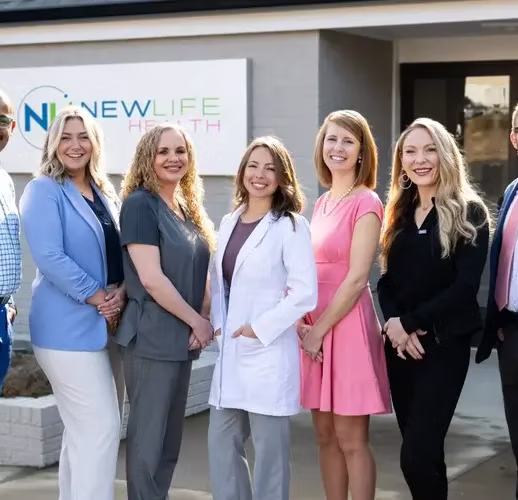 Seven people, including medical staff in professional attire, stand smiling outside a building with a "New Life Health" sign.