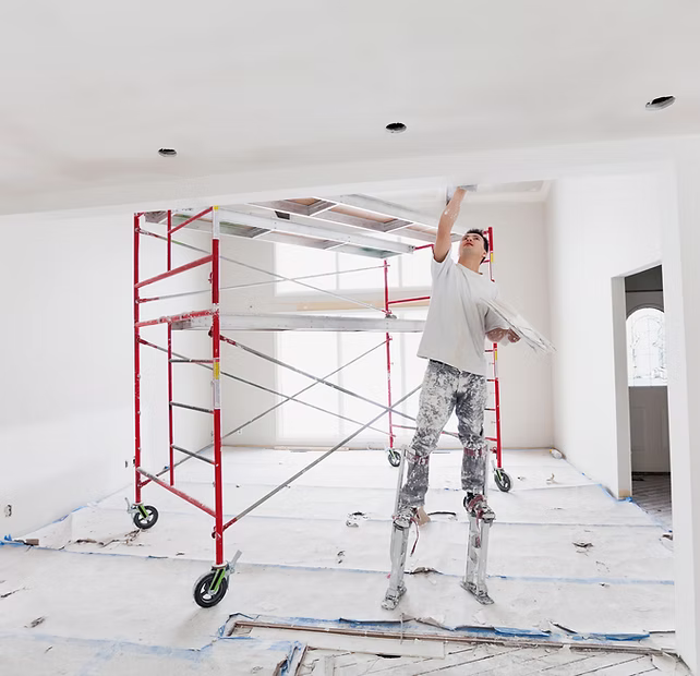 A worker on stilts paints a white ceiling in a room with scaffolding and protective floor covering.