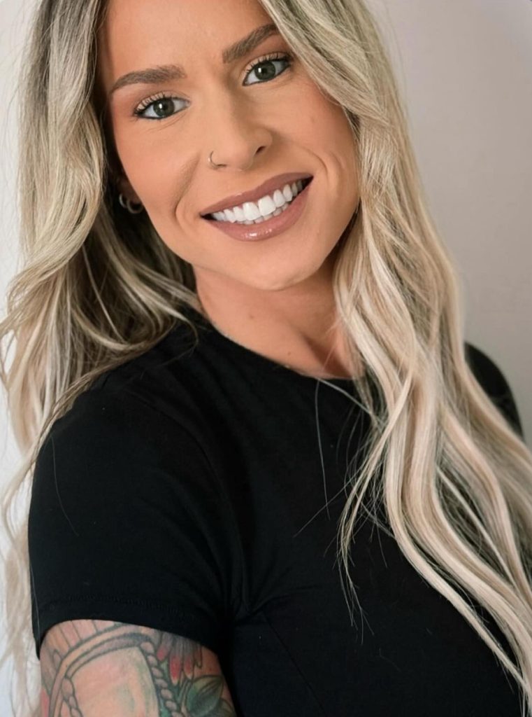 Smiling person with long blonde hair, wearing a black shirt, showing a tattooed arm.