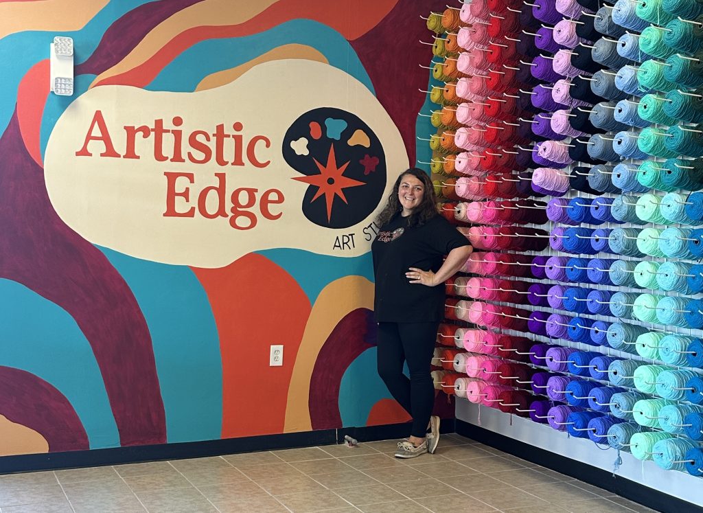 Woman stands by a colorful wall mural and shelves of yarn at Artistic Edge Art Studio.