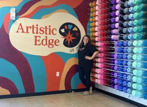 Woman stands by a colorful wall mural and shelves of yarn at Artistic Edge Art Studio.