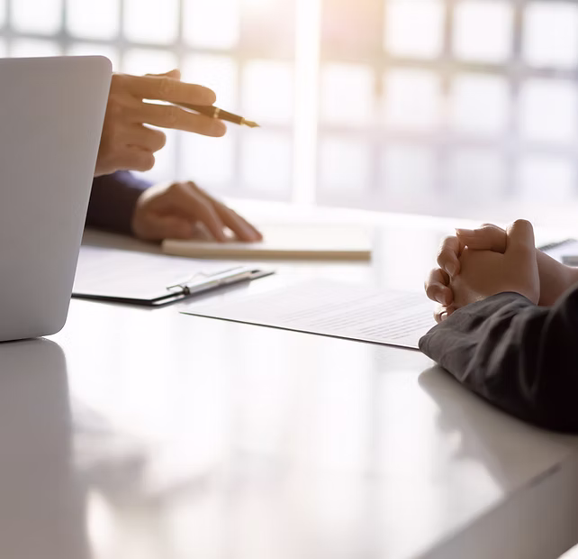 Two people sit at a desk with papers and a laptop, one gesturing with a pen during a discussion.