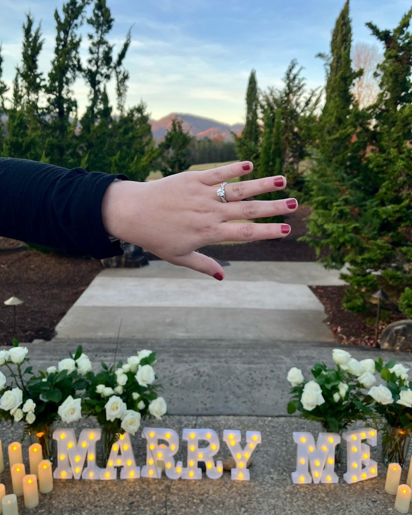 A hand with an engagement ring is held out above "MARRY ME" lights, flowers, and candles outdoors.