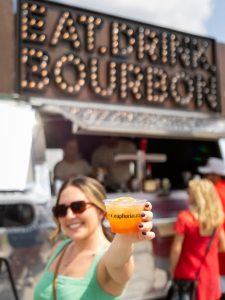 Smiling woman holding a drink in front of a food truck with a lit "EAT. DRINK. BOURBON." sign above.