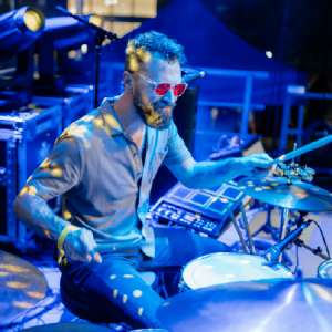 Man with red sunglasses playing drums on a stage under blue and yellow lights.