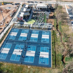 Aerial view of multiple blue pickleball courts with people playing, next to a building and outdoor seating area.
