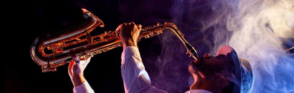 Man in a hat playing saxophone with dramatic lighting and smoke swirling around him.