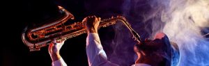 Man in a hat playing saxophone with dramatic lighting and smoke swirling around him.