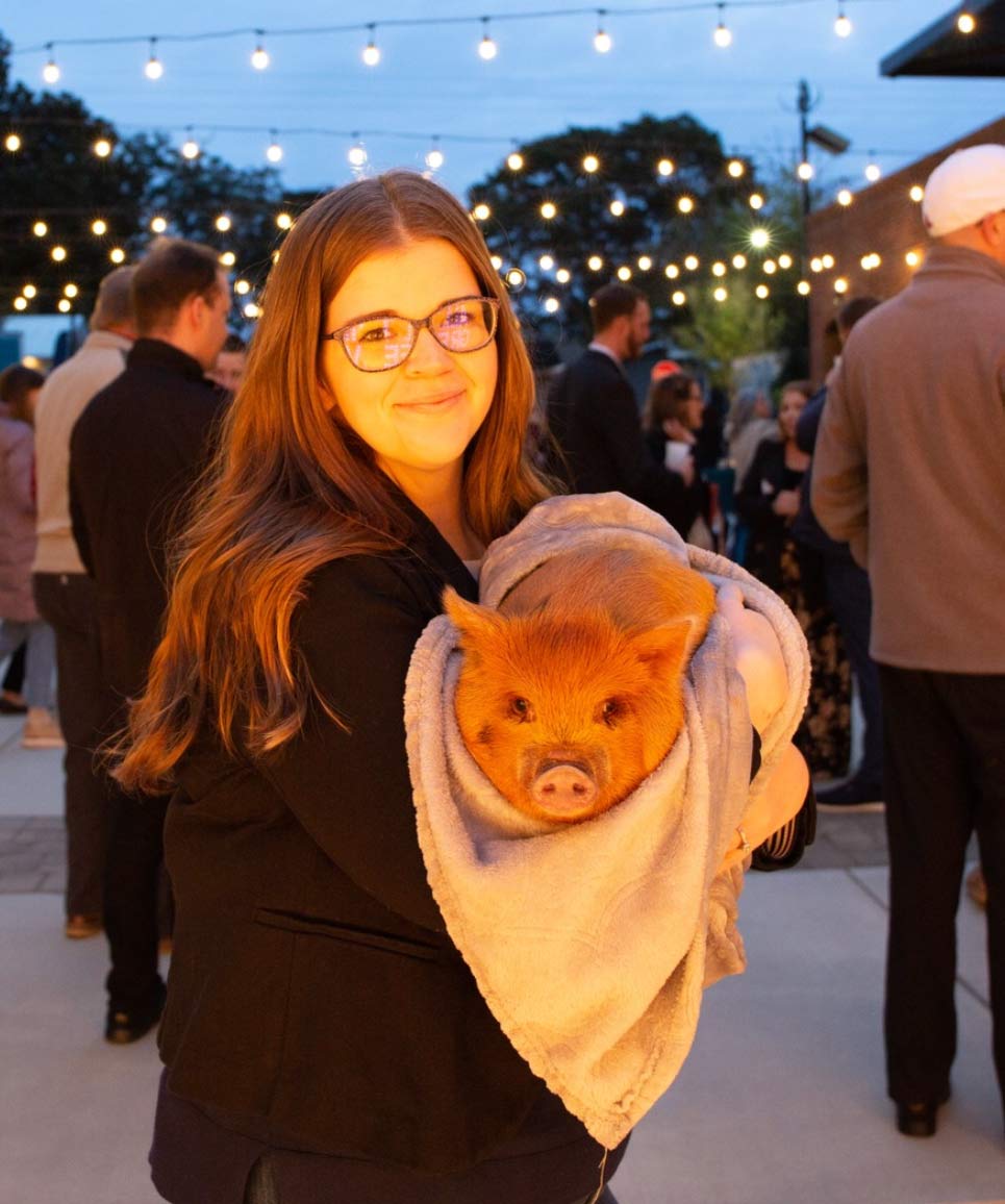 Woman with glasses smiling and holding a small pig wrapped in a blanket at an outdoor event with string lights.