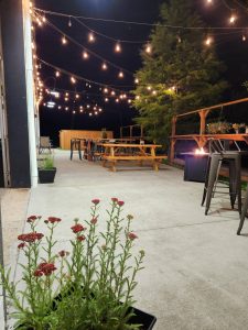 Outdoor patio at night with string lights, picnic tables, potted plants, and a fire pit.