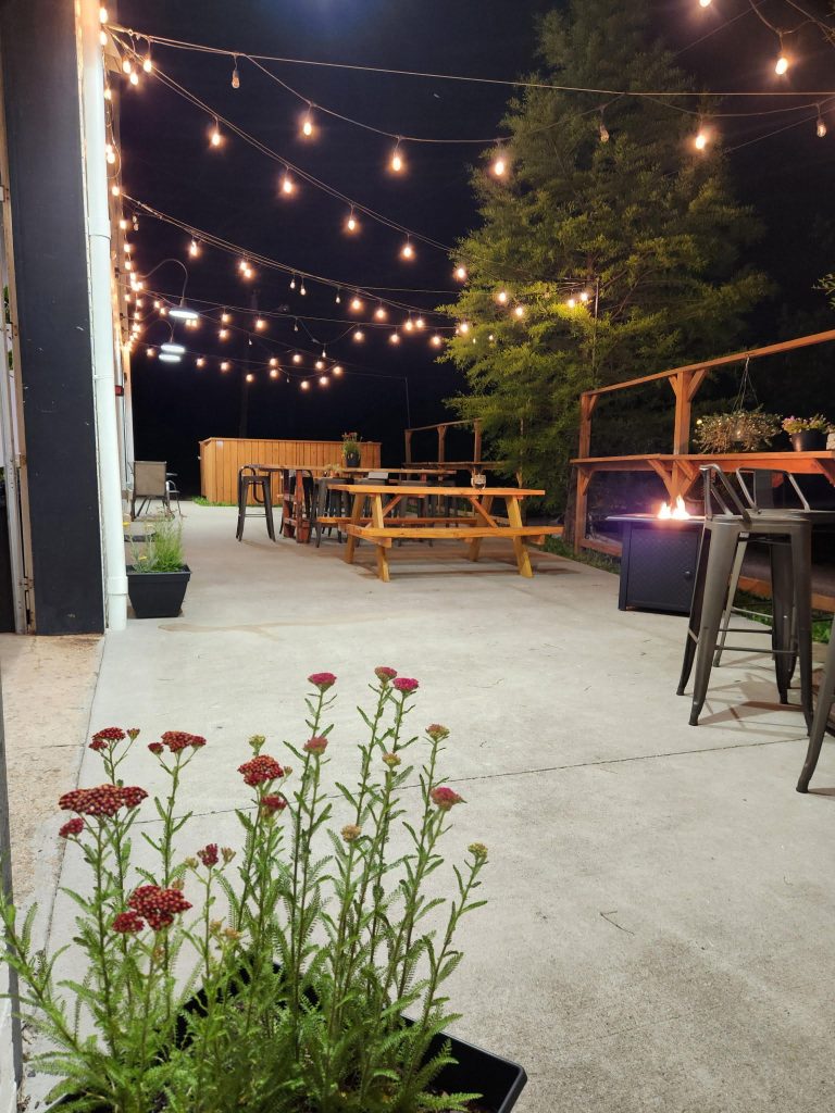 Outdoor patio at night with string lights, picnic tables, potted plants, and a fire pit.