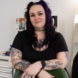 Woman with long dark hair, tattoos, and a black shirt smiling while sitting in a green chair in a bright room.