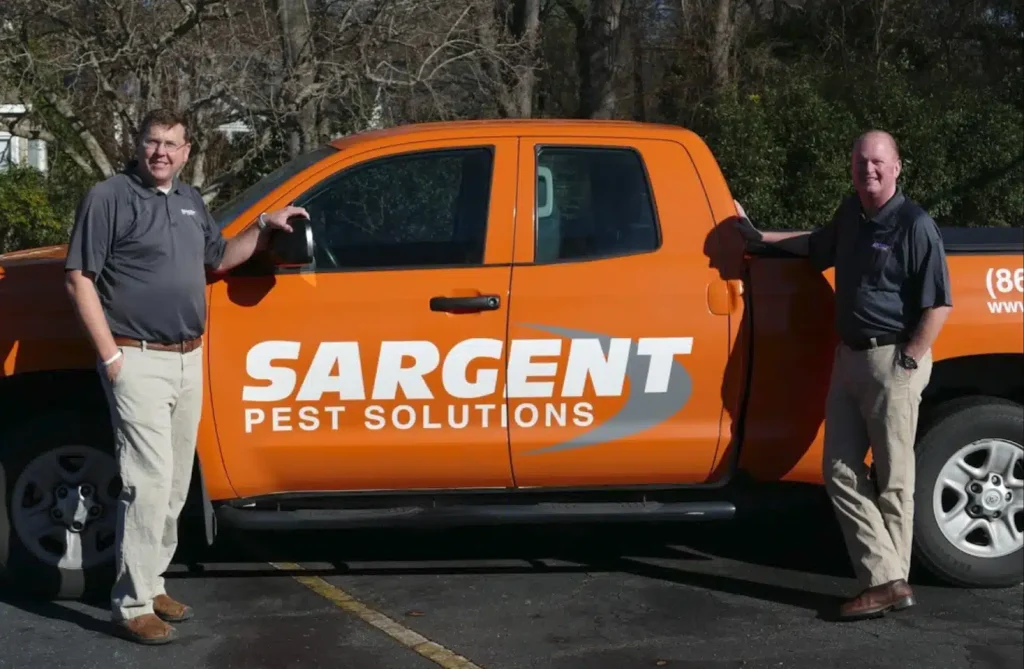 Two men stand by an orange Sargent Pest Solutions truck in a parking lot on a sunny day.