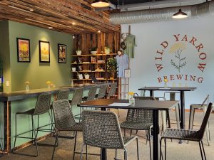 Modern brewery taproom with woven chairs, a bar, wall art, plants, and "Wild Yarrow Brewing" mural.