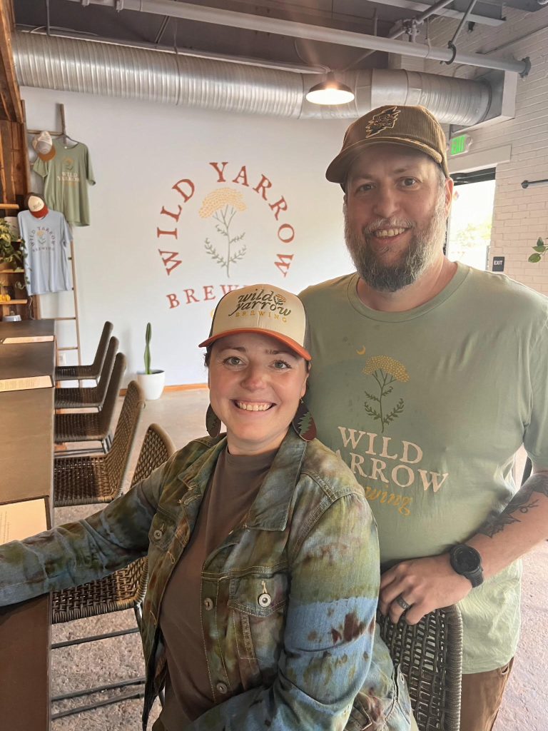 Two smiling people in Wild Yarrow Brewing shirts and hats stand inside the brewery near the logo on the wall.