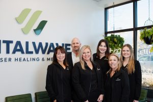 Six smiling staff members pose together in a modern medical office with the "VITALWAVE" logo on the wall.