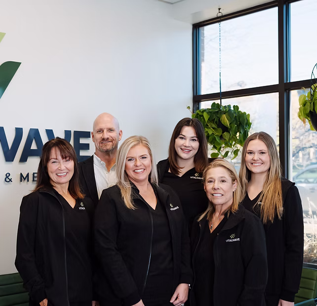 Six people in black uniforms smiling and posing together inside a bright office with plants and large windows.