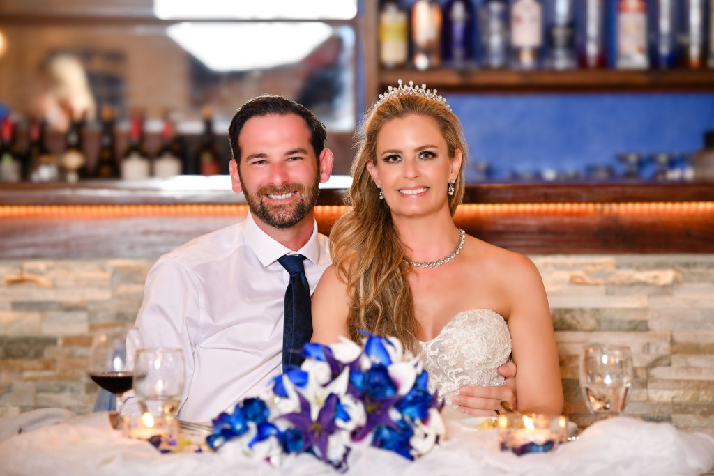 A bride and groom sit smiling together at a table with flowers, candles, and drinks in front of them.