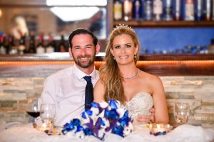 A bride and groom sit smiling together at a table with flowers, candles, and drinks in front of them.