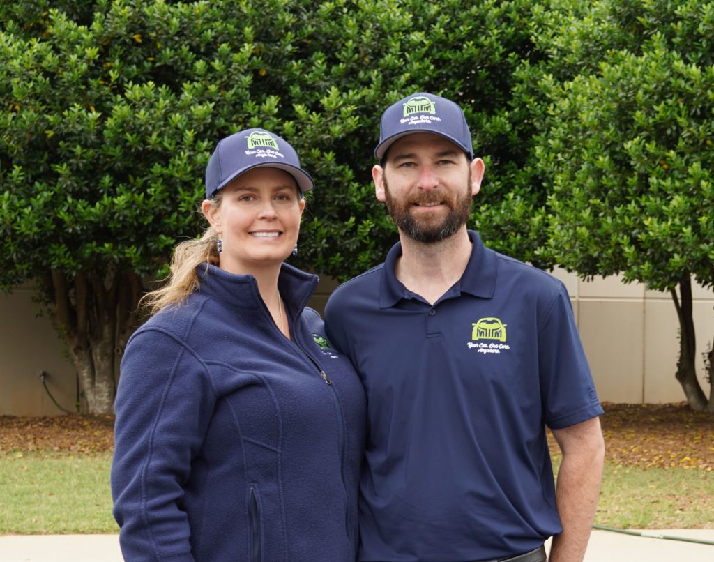 Two people in matching blue uniforms and hats stand outdoors, smiling in front of green trees.